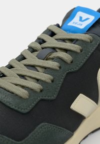 Sneaker featuring a dark green and black textile upper with grey laces and a blue logo tab. Light tan accent on the side.