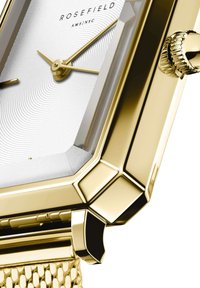 Reloj rectangular en tono dorado con una correa de malla texturizada, esfera blanca con patrón ondulado y manecillas elegantes en dorado. Presenta una corona texturizada.