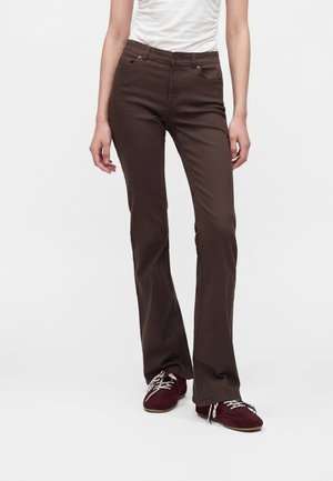 Femme portant un pantalon bootcut marron foncé, un haut plissé blanc et des baskets bordeaux avec des lacets blancs, debout devant un fond uni.
