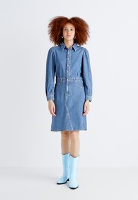 Vestido de ganga com gola, fecho de botões na frente, mangas longas bufantes, cintura cinto e saia evasê. Botas de cowboy azul claro.