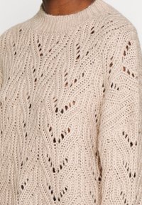 Gebreide trui in lichtbeige met een ajourpatroon, voorzien van een ronde hals en gestructureerd garen, met golvende motieven en openingen.