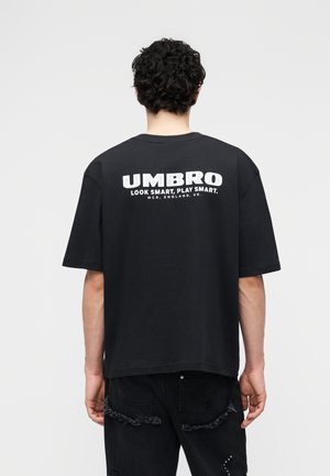 Person mit kurzen lockigen Haaren, der ein schwarzes, übergroßes T-Shirt trägt, auf dessen Rückseite in Weiß der Text "UMBRO Look Smart, Play Smart" steht.