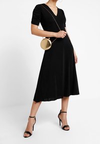 Sort knælængde kjole med korte puffærmer og V-udskæring. Sammen med en rund guldfarvet håndtaske og sorte strophæle.