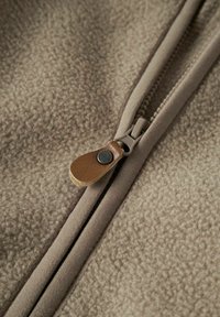 Primo piano di un tessuto in pile beige con una zip in pelle marrone e una zip di un marrone chiaro parzialmente chiusa.