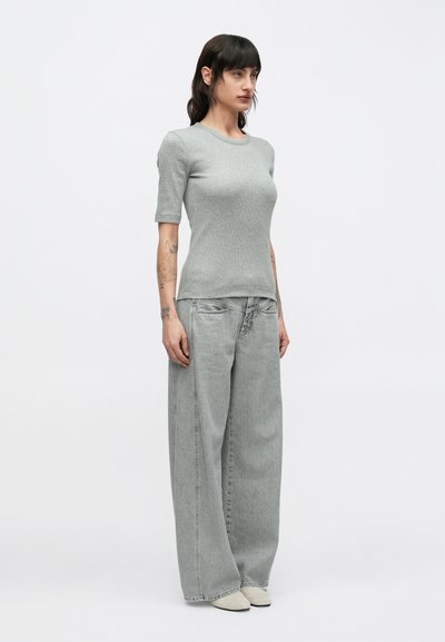 Γκρι ριμπ κοντομάνικη μπλούζα συνδυασμένη με ανοιχτόχρωμο γκρι τζιν παντελόνι σε άνετη γραμμή. Το μοντέλο φοράει ανοιχτό γκρι παπούτσια, στέκεται μπροστά σε απλό φόντο.