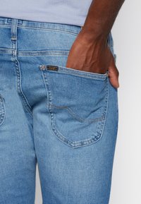 Mörkhyad hand placerad i bakfickan på ljusblå Lee-jeans som bärs med en ljusfärgad skjorta.