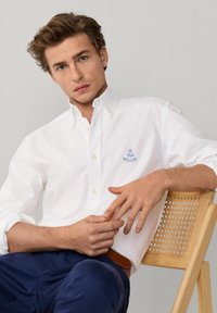 Giovane uomo con capelli castano chiaro che indossa una camicia bianca con bottoni e pantaloni blu navy, seduto su una sedia di legno con schienale intrecciato.