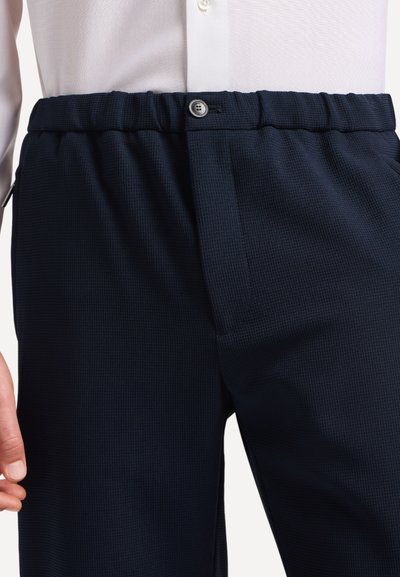 Primo piano di una persona che indossa pantaloni navy in tessuto elasticizzato con vita a elastico, abbinati a una camicia bianca con bottoni.