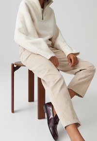 Personne assise sur un tabouret en bois portant un pull polaire crème zippé, un pantalon beige et des chaussures en cuir marron foncé sans lacets.