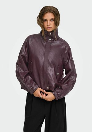 Giovane donna con capelli mossi che indossa una giacca di pelle viola scuro lucida con collo alto e tasche con bottoni automatici e pantaloni neri, in piedi con le mani giunte.