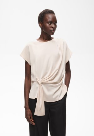 Femme aux cheveux courts portant un chemisier beige à manches courtes avec un nœud sur le devant et un pantalon noir, debout devant un fond blanc uni.