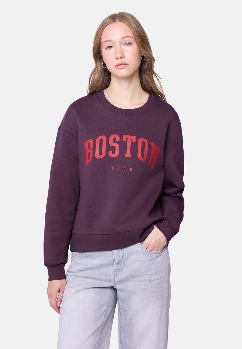 Fioletowa bluza z napisem "BOSTON" w pogrubionej czerwonej czcionce oraz rokiem "1980" poniżej. Posiada okrągły dekolt i ściągacze, zestawiona z jasnoszarymi spodniami.