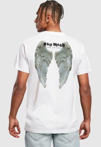 T-shirt en coton blanc avec un graphisme d'ailes d'ange en gris au dos, avec "Sky High and Beyond" imprimé au-dessus en gras noir.