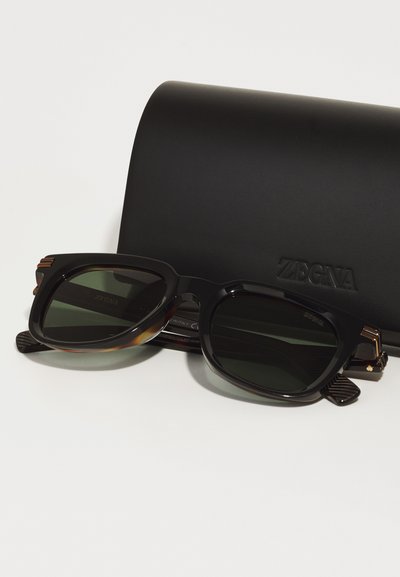 Black sunglasses with rounded edges, green lenses, and textured black arms; accompanied by a black hard case embossed with "ZEGNA."