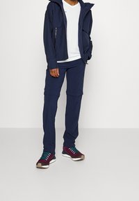 Persona vistiendo una chaqueta de cremallera azul marino y pantalones sobre una camiseta blanca, con zapatillas burdeos y verde azulado, de pie contra un fondo liso.