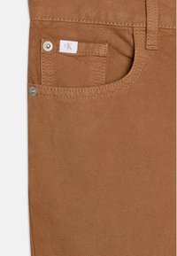 Pantalones de algodón marrones con un acabado texturizado, que presentan un pequeño bolsillo frontal, una etiqueta con el logo de CK y un detalle de botón metálico.