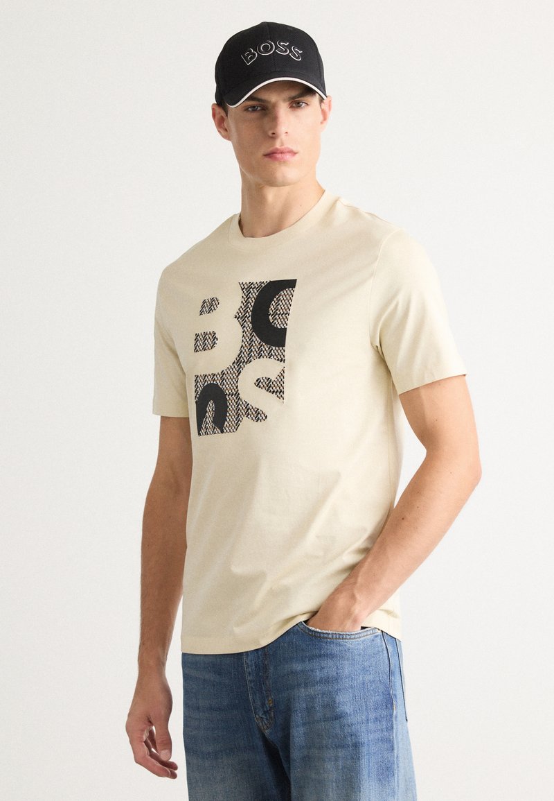 Man wearing beige BOSS logo t-shirt, blue jeans, and black BOSS cap, standing with one hand in pocket against plain background.