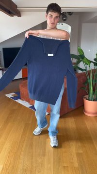 Mladý muž drží tmavě modrý svetr před sebou v moderním obývacím pokoji s dřevěnou podlahou a rostlinami.