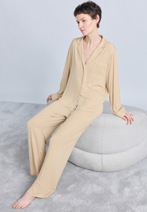 Γυναίκα με κοντά μαλλιά, ντυμένη με μπεζ ρούχα χαλάρωσης, κάθεται ξυπόλητη σε ένα υφασμάτινο στρογγυλό μαξιλάρι πάνω σε γκρι χαλί.