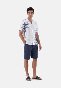 Kurzärmliges weißes Leinenhemd mit blauem Blattmuster, kombiniert mit navyblauen Leinen-Shorts und schwarzen Sandalen, vor einem schlichten Hintergrund stehend.