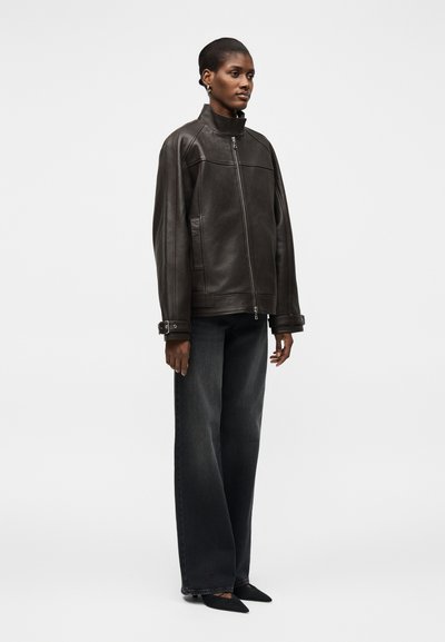 Γυναίκα με υπερμεγέθη μαύρο δερμάτινο μπουφάν με φερμουάρ και μανσέτες με αγκράφες, συνδυασμένο με φαρδιά σκούρα τζιν και μαύρα μυτερά παπούτσια, όρθια πλάγια.