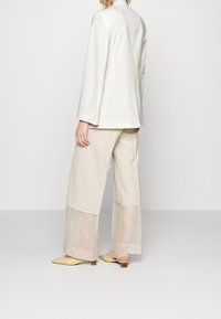 Beżowa dopasowana marynarka z długimi rękawami i guzikami, zestawiona z beżowymi spodniami o szerokich nogawkach oraz żółtymi mulesami na niskim obcasie z zaostrzonym noskiem.