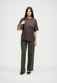 Hnědé oversized tričko, volné olivově zelené kalhoty a černé platformové boty. Drží malé černé kabelky. Jednoduchý design s minimálními akcenty.