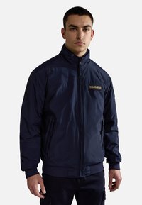 Mörkblå zip-up-jacka gjord av lättviktsmaterial, med hög krage, två sidofickor och en diskret logodetalj på bröstet.