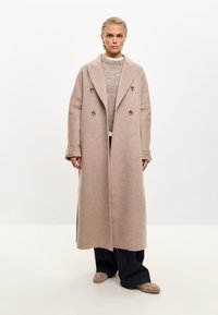 Cappotto doppiopetto in misto lana beige con ampi rever e bottoni di legno, indossato sopra un leggero maglione a maglia e pantaloni larghi scuri.