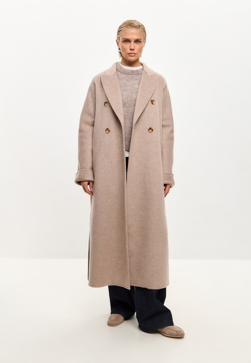 Abrigo de mezcla de lana beige de doble botonadura con grandes solapas y botones de madera, llevado sobre un suéter de punto ligero y pantalones oscuros de pierna ancha.