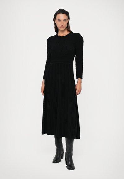 Vestido preto com textura canelada, mangas longas, com um corpete justo e uma saia rodada. Usado com botas altas de salto preto.