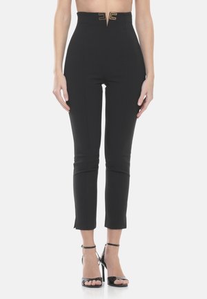 Donna che indossa pantaloni alla caviglia neri aderenti a vita alta con dettaglio fibbia dorata, abbinati a sandali neri con tacco alto e punta aperta.