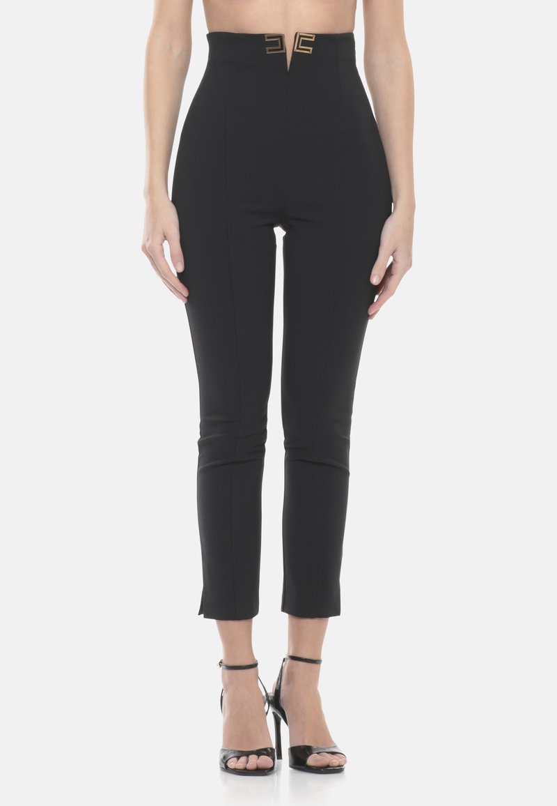 Frau trägt hoch taillierte, schwarze, schmal geschnittene Knöchellange Hosen mit einem goldenen Schnallendetail, kombiniert mit schwarzen, offenen High-Heel-Sandalen.