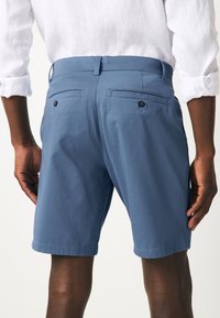 Modri bombažni kratke hlače z gladko teksturo, dvema zadnjima žepoma z gumbi in sproščenim krojem. Robovi so urejeni in ravni.