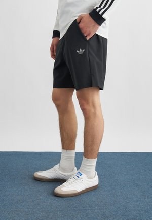 Person iført sorte Adidas-shorts med logo, hvid langærmet top med sorte striber, hvide sokker og hvide Adidas-sneakers på blåt tæppe.