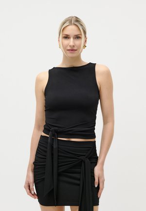 Blonde vrouw die een mouwloze zwarte top en een zwarte rok met gestrikte stofdetails draagt, staand tegen een effen lichte achtergrond.