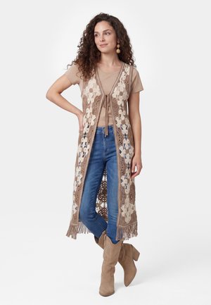 Kvinde med krøllet hår iført brune støvler, blå jeans, en beige t-shirt og en lang beige og cremefarvet hæklede vest med frynser, stående med den ene hånd på hoften.