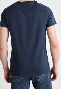 Uomo che indossa una camicia a maniche corte blu navy semplice e jeans blu, girato di spalle rispetto alla fotocamera su uno sfondo bianco.