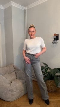 Bela kratka majica s kratkimi rokavi združena z visokim pasom sivih kavbojk. Črni škornji s peto, sedeči črtasti blazina v ozadju in notranje rastline.