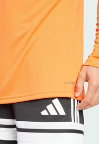 Oranges longärmliges Sportshirt mit glatter Textur und 'AEROREADY'-Logo nahe dem Saum, kombiniert mit schwarzen Shorts, die weiße Streifen und Logo aufweisen.