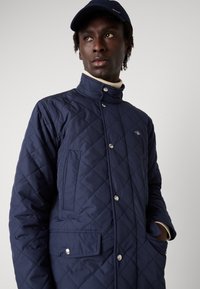 Tmavě modrá prošívaná bunda s vysokým límcem, zapínáním na patentní knoflíky a bočními kapsami; s jemným logem na hrudi a texturovanou látkou.