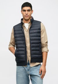 Ναυτικό γιλέκο με καπιτονέ σχέδιο, ψηλό γιακά, μπροστά με φερμουάρ και ρίγες. Φορεμένο πάνω από μια μπεζ μπλούζα και μπλε τζιν.