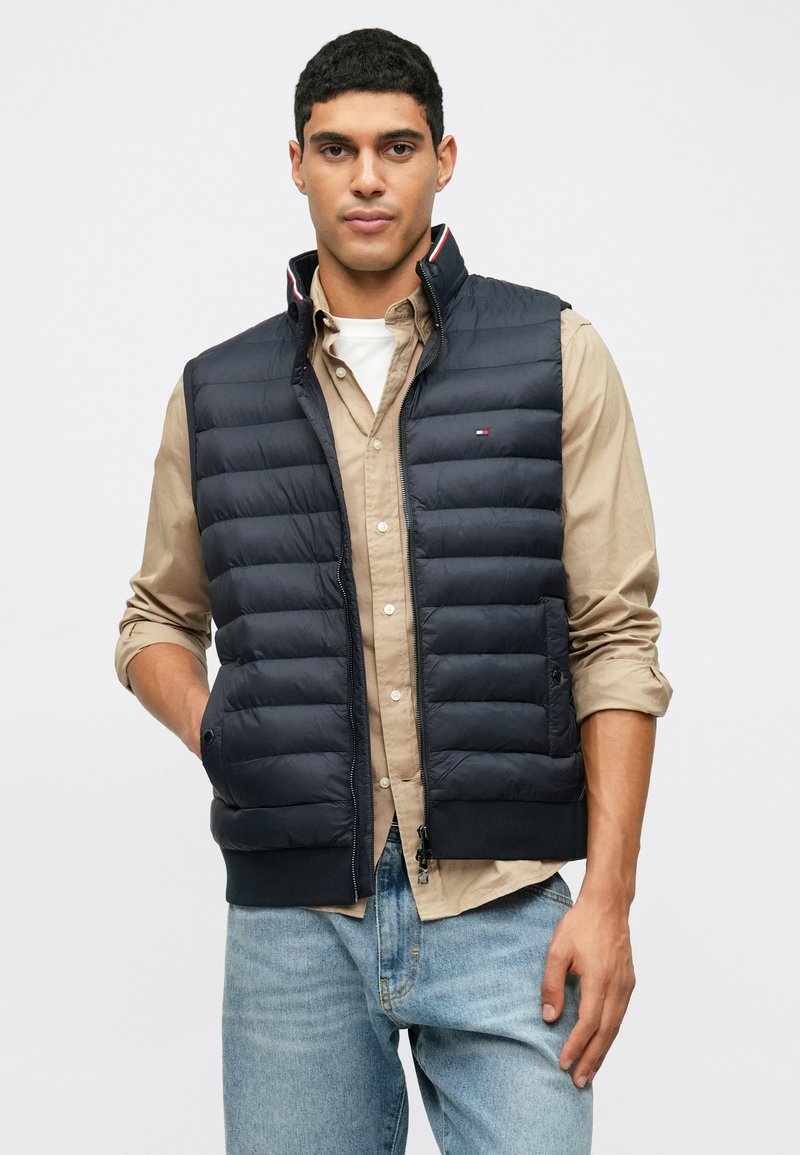 Marineblauer Steppweste mit gestepptem Design, hohem Kragen, Frontreißverschluss und gestreiften Akzenten. Über einem beigen Hemd und blauen Jeans getragen.