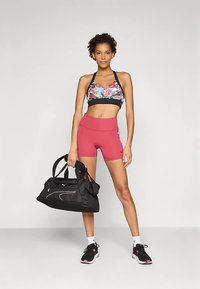 Conjunto deportivo compuesto por un sujetador deportivo multicolor, pantalones cortos rojos y zapatillas negras. En una mano sostiene una bolsa de deporte negra con detalles blancos.
