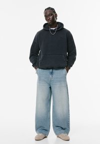 Schwarzer Kapuzenpulli mit Fronttasche, kombiniert mit hellblauen weit geschnittenen Jeans und beigen Sneakers. Das Model steht mit Händen in den Taschen.
