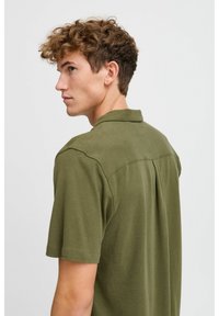 Camisa de manga corta en tela verde oliva, con cuello, detalles de costura y un diseño de pliegue en la parte trasera, que ofrece un ajuste relajado.