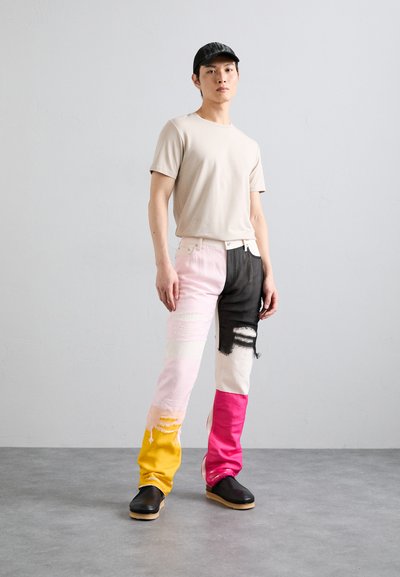 T-shirt beige a girocollo abbinato a jeans a toppa multicolore in rosa, nero, giallo e bianco con dettagli strappati. Indossati zoccoli neri.