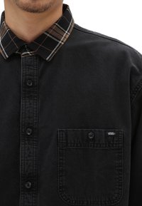 Uomo che indossa una camicia nera con bottoni e colletto a quadri, con tasca sul petto abbottonata e un piccolo tag con il logo Vans.
