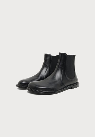 Paire de bottines Chelsea en cuir noir avec panneaux latéraux élastiques et semelles plates sur un fond clair uni.