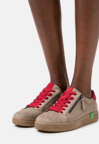 Sneaker in suede beige con lacci rossi, cerniera laterale e accento del logo verde. Presenta una spessa suola in gomma marrone e dettagli cuciti.